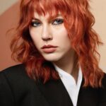 Person with wavy, shoulder-length red hair and bangs, wearing a black blazer over a white shirt, looking directly at the camera against a neutral background.