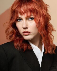 Person with wavy, shoulder-length red hair and bangs, wearing a black blazer over a white shirt, looking directly at the camera against a neutral background.