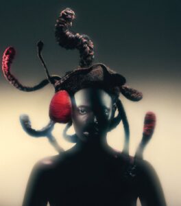 A person with dark skin is wearing an elaborate headpiece featuring thick, textured, tentacle-like forms, some wrapped with red yarn, against a gradient background.