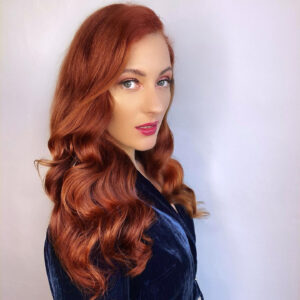 Woman with long, wavy red hair and light eyes wearing a dark blue velvet top, posing against a plain light background.