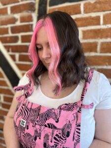 A woman with shoulder-length wavy brown hair featuring bright pink front sections, wearing a white shirt and pink zebra-print overalls, stands in front of a brick wall.