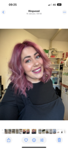 Woman with shoulder-length wavy pink hair and a black top smiles at the camera in a room with shelves and household items behind her.