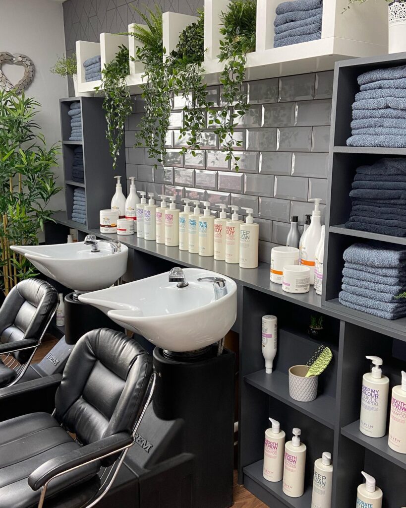 A hair salon wash station with two black chairs, two white sinks, shelves stocked with towels and various hair care products from Scissors of Eastney. Green plants decorate the space, and gray subway tiles line the wall.